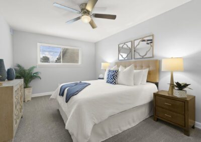 A bedroom with a ceiling fan and a neatly made bed, side table with lamp and table with vases and plant pot on the side